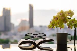 Spectacles put on open book with a cup of coffee, phalaenopsis orchid and city background. photo