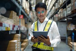 Portrait of a happy Asian man factory manager using a digital tablet in a warehouse. Inspecting goods and materials on shelves with inventory background logistics and export photo