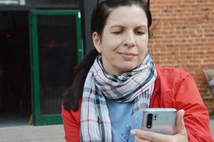 A woman in a red jacket, a light scarf and sunglasses reads a message, looks at something in a smartphone, sitting on a street bench. Correspondence, online learning, communication in social networks. photo