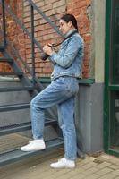 A young woman photographer in a blue denim jacket and glasses stands against the background of a brick wall, with a camera in her hands and looks through photos. Girl with a camera. photo