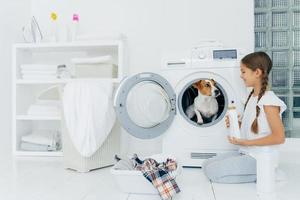 Positive small girl has fun with jack russel terrier, poses on floor near washing machine in laundry room, holds liquid powder, busy with housework, has glad expression. Cozy interior in bathroom