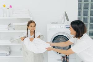 Happy woman with dark short hair pulls off laundry from basket, happy child poses in it, spend time in bathroom near washing machine and iron on top, shelf with folded white towels. Laundry time photo