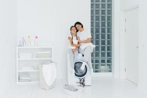 Happy mother and daughter have fun, embrace and express love to each other, dressed in casual wear, sit on washer, prepare for washing, being in white spacious laundry room, basin with dirty clothes photo