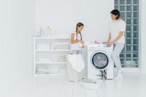 Indoor shot of happy mother and daughter stand near washing machine, girl pours liquid powder, load washer with dirty clothes, do housework, have laundry day at home. Household chores concept photo