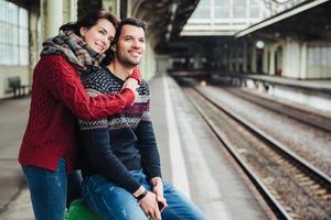 Pleasant looking female and male embrace each other stand near luggage at platform, going to say goodbye each other and have seperation, look pensively into distance. Travelling and vacation photo