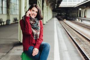 Beautiful brunette female wears knitted sweater and scarf, sits on suitcase near platform, communicates on smart phone, feels bored to wait for long time. People, travelling and waiting concept photo