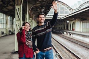 Lovely couple being on platform of railway station, wave someone as look into distance, meet their relatives or friends. Young married woman and man say goodbye someone. Farewell concept. photo