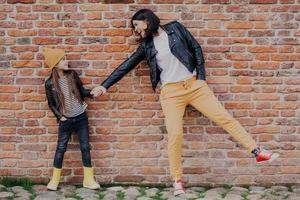 la pequeña niña de moda y su madre se divierten juntas, mantienen las manos juntas, posan contra la pared de ladrillos. elegante joven madre e hija juegan al aire libre. relaciones, buen humor y concepto de moda. foto