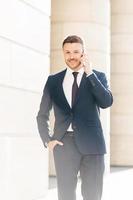 Vertical shot of handsome successful male financial director solves problems via smart phone, looks confidently at camera, keeps hand in pocket, poses outdoor. People and technology concept. photo