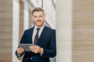 Confident cheerful male lawyer reads business news, has gentle smile, dressed in formal clothes, poses in urban setting. Businessman checks email or updates profile on digital tablet computer photo