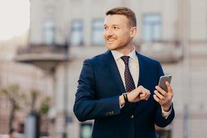 Horizontal shot of successful unshaven male CEO looks for interesting multimedia files on smart phone, downloads or updates new programs, stands against blurred city background, looks happily aside photo
