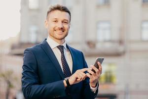 Satisfied businessman holds smart phone, reads email from investor, happy to recieve good news about business company, stands in urban territory, wears black suit and elegant tie. Technology concept photo