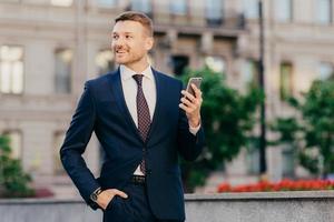 Happy attractive male financier in elegant wear, uses modern smartphone for following financial news on special installed application, stands on street, looks aside with cheerful expression. photo
