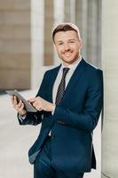 Vertical shot of business person uses digital tablet for online communication and work, uses free internet at office, has positive expression, dressed in formal black suit. People and occupation photo