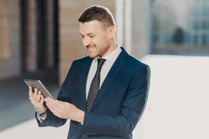 Cheerful male manager reads notification on digital tablet, wears formal black suit with tie, connected to wireless internet, watches video on web page. Business and modern technologies concept photo