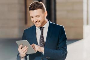 Confident handsome male manager checks email on tablet computer, updates database on touch pad, wears elegant formal clothing, connected to wireless internet, poses outdoor. Business concept photo