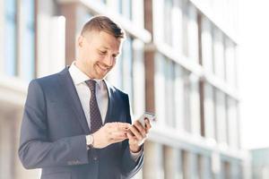 Young prosperous businessman chats online or reads information via cell phone, connected to wireless internet, poses against urban background outdoor, has happy expression. People and wok concept photo