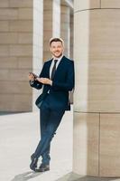 Photo of cheerful male dressed in expensive black suit, stands crossed legs, works on touch pad, stands in modern office interior, smiles positively at camera. Young male manager. Full length portrait