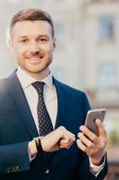 Smiling manager director with watch on arm, dressed elegantly, holds smart phone, reads positive news about his new project, poses outdoor, waits for colleague near office building. Job concept photo