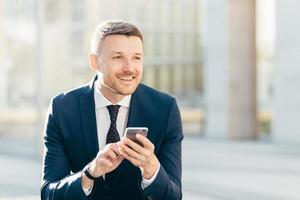 Delighted thoughtful elegant male marketing trader in formal black suit, uses modern cell phone for searching website in internet, types text message, looks pensively into distance. Bussiness concept photo