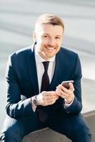 Positive successful male CEO sits outdoor, uses modern smart phone for reading news in message, checks notification, dressed in elegant suit, has satisfied expression, enjoys online communication photo