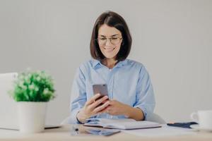 Happy female employee works on developing new project in office, searches information on websites via modern electronic gadgets, uses free internet connection in her cabinet, has happy expression photo