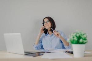 Attractive brunette female journalist arranges meeting via cell phone, works on new article on laptop computer, holds pen in hand as going to write some details in her notebook. Busy worker in office photo