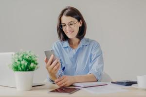 Pretty successful talented female copywriter blogs on smart phone, works on laptop computer, checks notification on modern electronic gadget, writes something in notebook with pen. Technology, job photo