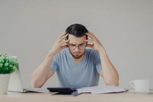 Brunet young male faces financial problems, not able to pay out mortagage, has desperate expression, makes calculations with calculator, isolated over grey background, uses modern technologies photo