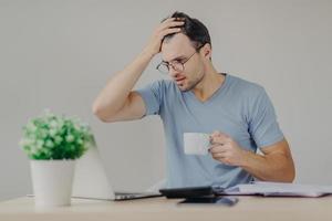 Young frustrated male does paperwork, reads with puzzled expression unpleasant information on laptop computer, drinks strong coffee, surrounded with calculator and documents on working table photo