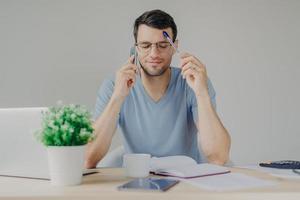 Busy male works on organizational issues, speaks with business partner via cell phone, keeps eyes closed as tries to remember necessary information and write in diary. People and work concept photo