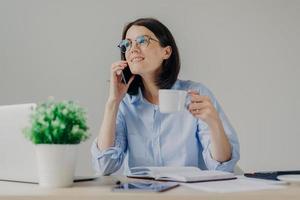 Gorgeous pleased businesswoman has cell phone conversation while has coffe break after working on notebook, shares her success with close person, isolated over grey background. Occupation concept photo