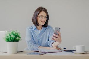 Successful brunette female freelancer recieves good news on smart phone while works on laptop computer, reads post on website, makes notes in diary, drinks strong coffee, makes some calculations photo