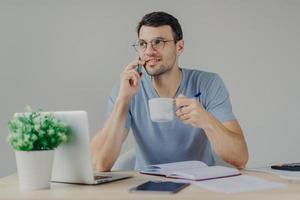 Pleasant looking male accountant in round glasses sips hot beverage, has mobile conversation, manages finances, works with laptop computer, writes notes in diary. Man answers call from bank. photo