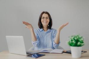 Happy overjoyed female achieves success in her work, going to present high results, raises hands joyfully, sits at working place with laptop computer, diary, calculator, isolated on grey background photo