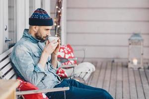 Thoughtful handsome male with beard and mustache, drinks hot tea, warms after stroll, wears fashionable clothing. Pensive man student plans how to pass all exams successfully. Calm atmosphere photo