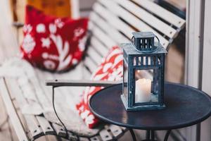 Shot of white candle with burning flame in lamp, stands on round table, bench with cushion in background. Cozy domestic interior. Wonderful decor photo