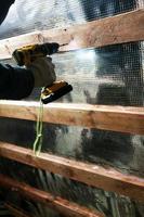 The wizard uses a cordless drill and screw the screws into boards that hold foil film and mineral wool to heal the roof of the house photo