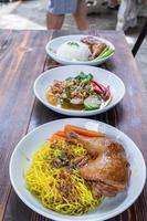Roasted chicken thighs and drumsticks are served with yellow noodles and fresh vegetables in a white plate. Chicken thighs baked in a sweet and delicious sauce are served with hot yellow noodles.