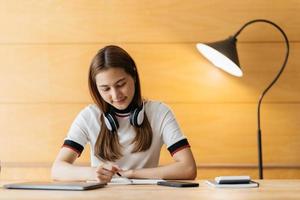 asiático feliz en el uso de auriculares portátil tomar cursos de capacitación en línea tomar notas. una joven estudiante asiática sonriente con auriculares estudia en una computadora desde casa. concepto de educación a distancia. foto