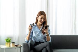 smiling asian woman holding smartphone and banking credit card, involved in online mobile shopping at home, happy female shopper purchasing goods or services in internet store. photo