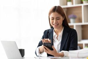 Asian businesswoman in formal suit in office happy and cheerful during using smartphone and working, copy space. photo