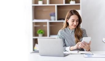 retrato de una mujer de negocios asiática que paga facturas en línea con una computadora portátil en la oficina. hermosa chica con computadora y chequera, feliz pagando facturas. concepto de cuenta de cálculo financiero de negocio de inicio foto