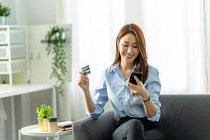 smiling asian woman holding smartphone and banking credit card, involved in online mobile shopping at home, happy female shopper purchasing goods or services in internet store. photo