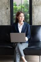 mujer asiática escribiendo correo electrónico en una computadora portátil mientras está sentada en casa, mujer de negocios joven que trabaja en una oficina moderna de inicio creativo. analizar acciones de mercado, nueva estrategia. foto