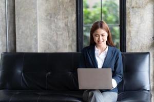 mujer asiática escribiendo correo electrónico en una computadora portátil mientras está sentada en casa, mujer de negocios joven que trabaja en una oficina moderna de inicio creativo. analizar acciones de mercado, nueva estrategia. foto