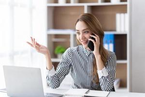 Young business woman on the phone at office. Business woman texting on the phone and working on laptop. Pretty young business woman sitting on workplace. Smiling business woman. photo