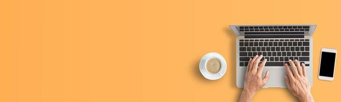 Person hand using laptop computer from above with coffee and smartphone on orange background.