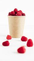 Fresh red raspberries in a paper cup on a white table background photo