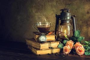 Coffee in cup glass on old books and clock vintage with kerosene lamp oil lantern burning with glow soft light on aged wood floor.